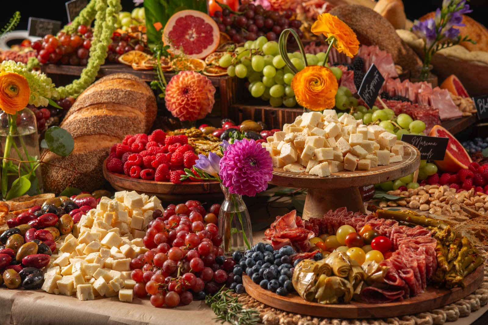 A lavish grazing table of cheeses, fruits, cured meats, breads, and flowers by Andiamo